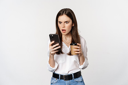 女人拿着咖啡杯看智能手机 震惊 惊吓脸部 站在白色背景上站立图片