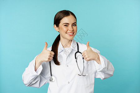 快乐的女医生 穿着白大衣的医务工作者 举起拇指来表示赞许 像某种东西 赞美 站在蓝背景上专家诊所实习生药品护士临床职业广告微笑看图片
