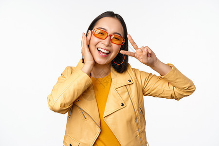 穿着太阳眼镜和黄色夹克 展示和平 V标志手势 站在白色背景上站立 笑笑脸的现代年轻女孩的肖像办公室成人促销工作室企业家太阳镜标识图片