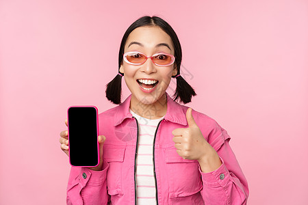 快乐时尚女孩推荐手机应用 微笑的亚洲女模特展示智能手机屏幕和大拇指 站在粉红色背景上情感互联网商业太阳镜促销细胞黑发标识学生快乐背景图片