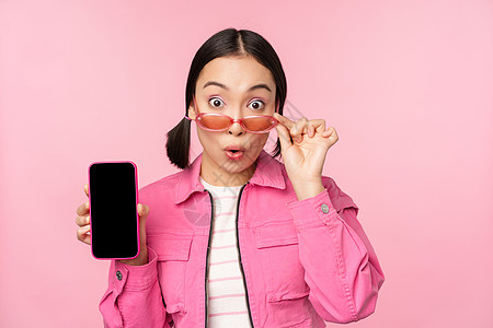 手机界面在粉红背景上站立 反应惊吓 感到惊讶 Asian女孩的肖像显示手机屏幕商业界面广告情感购物女朋友情绪大学学生标识背景