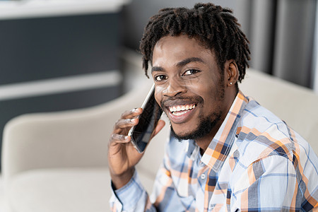 年轻的非洲裔美国人在谈话时手里拿着手机看着相机 坐在沙发上进行当前对话的快乐男人的画像 社交媒体概念图片