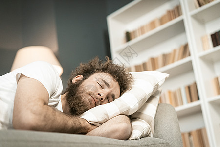 长时间工作到深夜后 睡在沙发上的留着胡子的英俊年轻人抱着枕头 生病的年轻人服药后睡在沙发上 远程工作概念图片