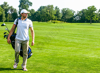 年轻的男高尔夫球手携带一袋高尔夫球杆 身穿白色 T 恤 高尔夫球鞋 白色帽子和米色裤子的苗条男子正在绿色草坪草丛中行走 复制空间图片