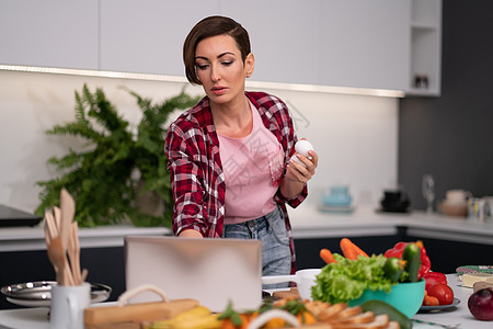 使用笔记本电脑搜索在线食谱的家庭主妇 同时用手做饭或烘烤鸡蛋 在厨房做饭的年轻女性 健康生活图片