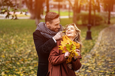 快乐的恋爱中的年轻人 男人从背后拥抱女人抚摸着她的脸颊 幸福的情侣穿着时髦的外套在秋天的公园里散步 捡起落叶 家庭和人的概念街道图片
