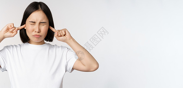 对亚洲女人的肖像 闭上耳朵和感到不适 从响声 烦人的声音 站立在白色背景上企业家工作室商业女朋友情绪衬衫女性学生成人技术图片