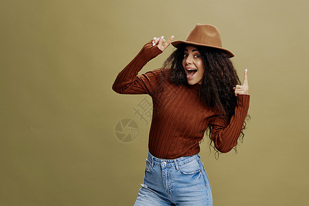 喜欢戴着棕色帽子的时尚卷发拉丁女性 对着镜头微笑 手牵着帽子 手指向上 孤立的绿色背景 复制空间服装时尚品牌 免费为您投放广告图片