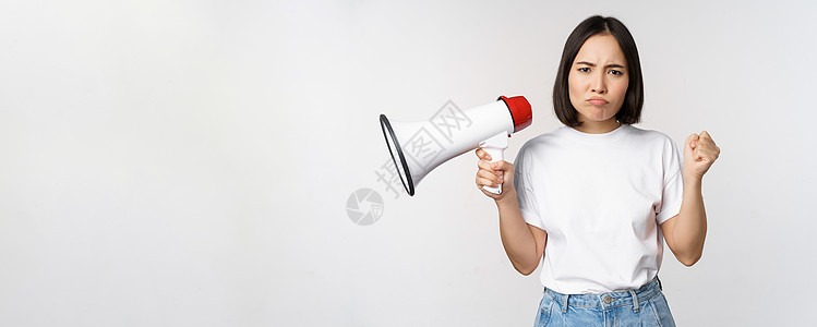 愤怒的亚洲女孩活动家 握着扩音机 看着愤怒 抗议 站在白背景上站立女士购物女性广告学生企业家商业互联网大学技术图片