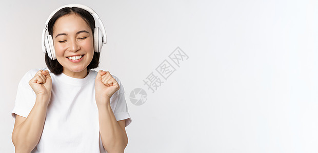 现代的亚洲女孩舞蹈 用耳机听音乐 笑笑快乐 站在白色背景的T恤衫上工作横幅情感商业学生广告成人情绪购物女性图片