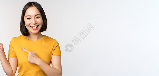 穿着黄色T恤衫微笑的亚裔黑发女孩的肖像 左手指向左 展示复制空间 Promo交易 示范旗帜 站在白背景上技术成人工作室女性商业女图片