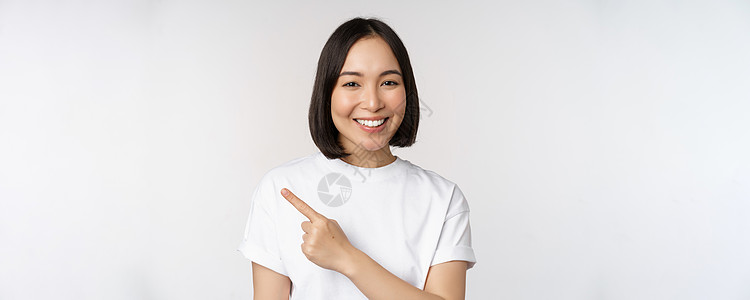 日本年轻女性的特写 她微笑着 手指向左指着复制空间 展示公告或广告横幅 站在白色背景上互联网情感学生情绪商业衬衫女士大学技术工作图片