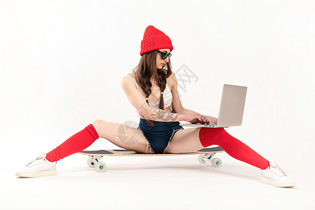 穿着红色帽子的女孩和红袜子坐在长板上 用笔记本电脑在白色背景上互联网发型木板性格滑板爱好运动技术快乐溜冰者图片