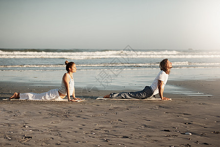 该地区的雄风会增加你的瑜伽 一对年轻夫妇在海滩上一起练瑜伽的镜头图片