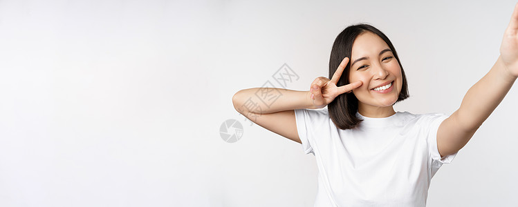 美丽的亚洲年轻女性自拍 用和平 V 字牌摆姿势 开心地笑着 拍照 在白色背景下摆姿势衬衫工作室技术学生女士女朋友企业家情绪成人大图片