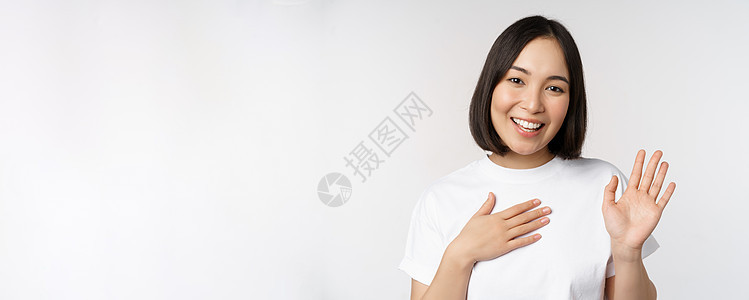 美丽的韩国女孩举手的画像 自我介绍 手放在心上 问候 站在白色背景上大学购物女朋友工作室情绪商业企业家横幅压痛技术背景图片