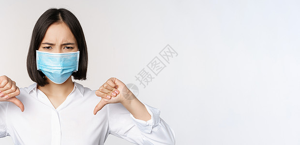 在医疗面罩上假面具的亚洲妇女脸部肖像显示拇指低下 表情失望 疲倦 站立在白色背景之上女士人士成人商业技术售货员面具女孩办公室冒充图片