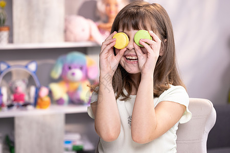 用蛋白杏仁饼干捂住眼睛的小女孩开心的笑脸 女学生玩甜点马卡龙 把饼干像眼镜一样放在眼睛周围 把手扔到两边图片