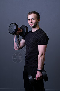 一个身穿黑色 T 恤 肌肉男背景 戴着哑铃的男人 中间的耳机对运动员的健康有好处 成人 t衬衫二头肌力量 健身房年轻图片