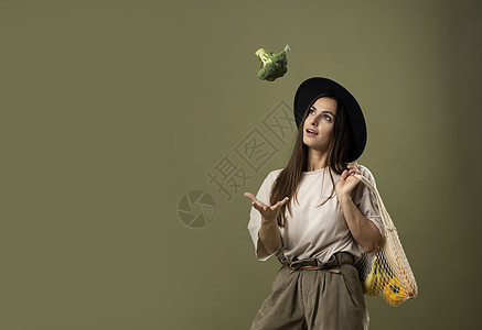 穿着米色 T 恤和帽子的黑发年轻女子微笑着 肩上拿着一个可重复使用的网袋 里面装着新鲜蔬菜和水果 并向空中扔西兰花 零废物概念图片