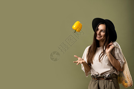 身穿美食T恤和黑帽子的生态友好笑容女性将黄胡椒单手扔在空气中 并肩上装满可再用棉皮果袋图片