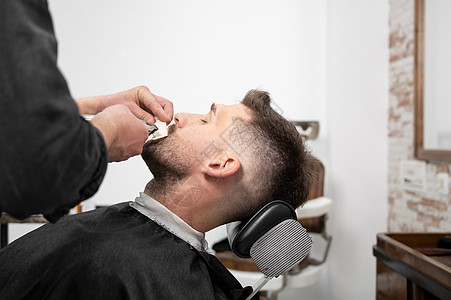年轻时髦的白人男子在现代理发店梳理胡须 男士发型设计 英俊的男人用电动修剪器换新发型剃须商业头发摄影剃须刀潮人顾客理发修剪剃刀背景图片