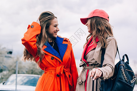 穿着橙色大衣 蓝色牛仔裤和领带的时尚快乐笑脸金发女子与街上女友沟通冒充金发女郎外套头发城市街道魅力奢华青年女性图片