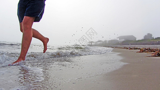 一个人在有雾的天气里沿着海滩奔跑的脚 海浪溅起的水花四处飞溅 穿着短裤奔跑的男人脚下的沙子和海浪 赤脚晨跑在沙滩上图片