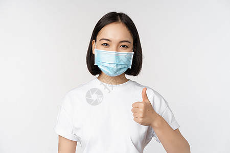 戴着医用面具微笑的亚洲女孩竖起大拇指 赞同一些好东西 赞美和赞美公司 站在白色背景上女性保健学生大学互联网购物卫生商业女士女朋友背景图片