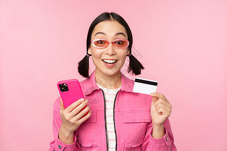 网上购物 微笑的亚洲女购物者 手持智能手机和信用卡 用移动应用支付 站在粉红色背景中学生太阳镜商业黑发卡片互联网女孩细胞信用电话图片