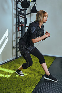 穿着传感器和腰带的漂亮金发女人 用维可牢尼龙搭扣固定 正在健身房练习 EMS 健身 做下蹲 现代运动重量俱乐部头发身体成人监控培图片
