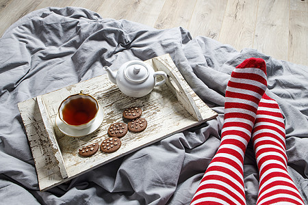 红色条纹袜子的腿片段 室内和家庭舒适的概念 顶视图 一杯茶 一壶凉茶 床上白色木制托盘上的糖碗 瓷杯图片