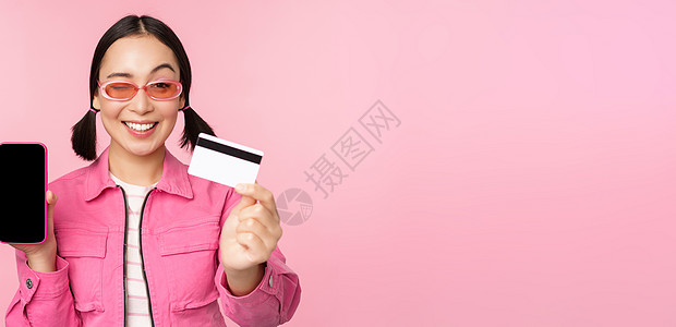 微笑的韩国女性展示信用卡和手机屏幕 智能手机应用程序界面 在线支付 非接触式购物 站在粉红色背景上的图像办公室女朋友促销电话商业图片