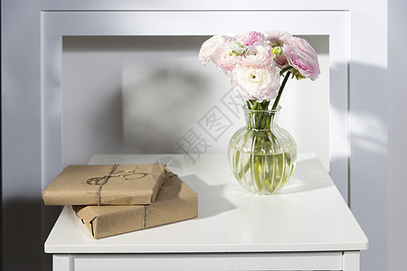 玻璃花瓶中的一束粉红色波斯毛茛和两件包装好的礼物放在假白色壁炉前的白色桌子上图片