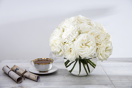 一束白玫瑰花 在一张桌边的圆玻璃花瓶里 加上一杯茶和一本书背景