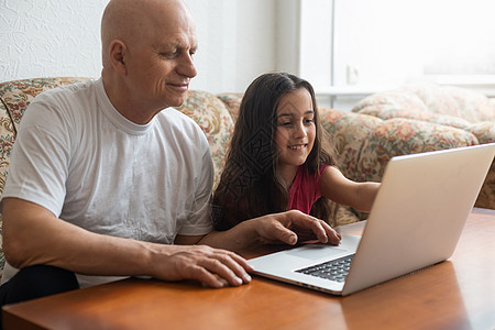 快乐的小女孩拥抱着微笑的祖父 坐在沙发上用笔记本电脑阅读爷爷互联网退休孙女学习青少年成人女孩头发图片