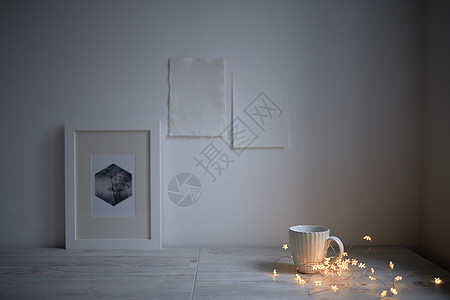 斯堪的纳维亚风格 室内设计 桌子上放着一个白色的杯子 一个舒适的发光花环灯 一个相框 两张白纸贴在墙上 文本的空白空间图片