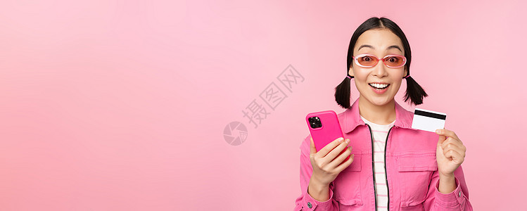 网上购物 微笑的亚洲女购物者 手持智能手机和信用卡 用移动应用支付 站在粉红色背景中黑发商业卡片工作室电话太阳镜女士女朋友广告促图片