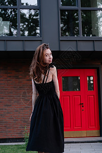 迷人的年轻女子穿着黑色连衣裙 站在带红门的现代建筑前 乔迁 买房子 私人住宅的幸福生活城市休息游客奢华黑发街道生活建筑学城镇人像图片