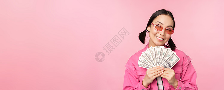 戴墨镜的漂亮韩国女人 展示美元 现金 微笑着高兴 快速贷款 小额信贷和支付的概念 站在粉红色背景上大学企业家快乐购物学生情感促销图片