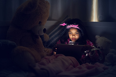 整晚都在娱乐 一个小女孩晚上躺在床上和她的泰迪熊一起使用数字平板电脑的裁剪镜头图片