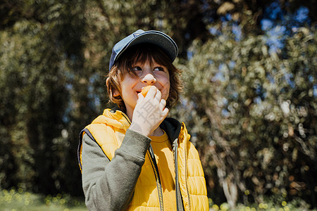 身穿黄色背心和绿色连帽衫的微笑快乐的孩子在公园户外吃清脆的小吃 男孩喜欢在外面吃嚼垃圾食品 背景是树木植被情感午餐油炸民众活力垃图片