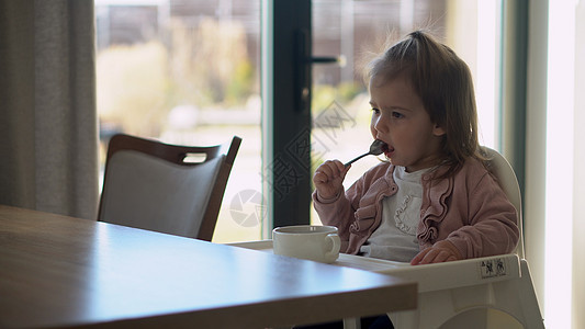 小婴儿坐在白色喂食高脚椅上的特写镜头 孩子正试图自己吃东西 快乐的孩子脸上沾满食物 小女孩用勺子吃粥 高品质的厨房早餐图片