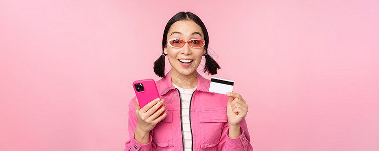 网上购物 微笑的亚洲女购物者 手持智能手机和信用卡 用移动应用支付 站在粉红色背景中卡片互联网女士电话标识促销信用商业成人办公室图片