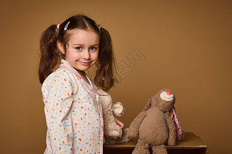 特写可爱的小女孩 有两条马尾辫 拿着她的毛绒玩具 自信地看着相机 可爱地微笑着 在米色背景中被隔离 有复制广告空间图片