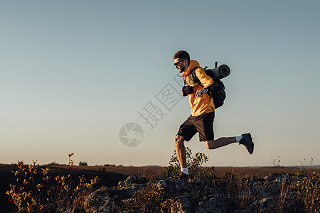 穿戴风速旅行男子服装 穿着背包和露营马甲 在日落时跳上山顶 男游牧人带数码相机享受他最好的独唱之旅图片