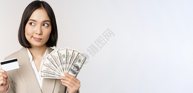 有思想的女商务人士 韩国女商务人士展示信用卡和现金 手中的美元 站在白人背景上思考商务信用工作室女性人士快乐售货员成人管理人员银图片