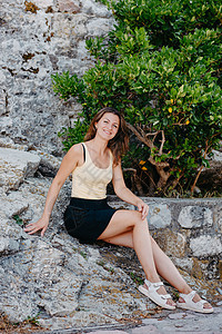 美丽的女孩坐在布德瓦老城的石墙上 女孩坐在石栏杆上 布德瓦 黑山旅游岩石堡垒城市女性裙子假期海滩微笑石头背景图片