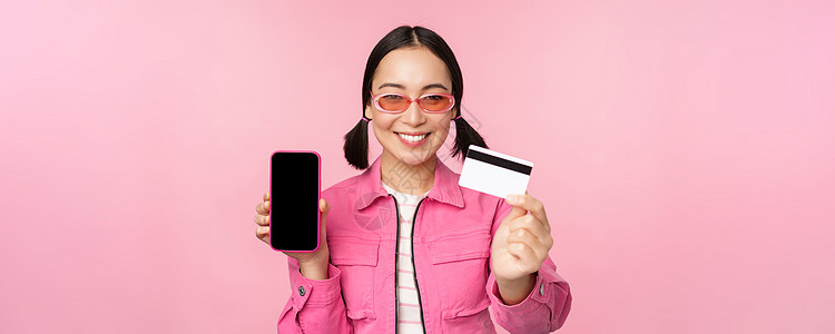 微笑的韩国女性展示信用卡和手机屏幕 智能手机应用程序界面 在线支付 非接触式购物 站在粉红色背景上的图像信用标识女士促销互联网商图片