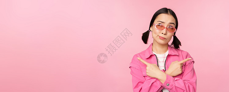 选择 时尚的韩国女孩 亚洲女模特手指侧身 展示两种变体 产品广告 展示物品 站在粉红色背景上情感困惑黑发太阳镜女士商业成人工作室背景图片